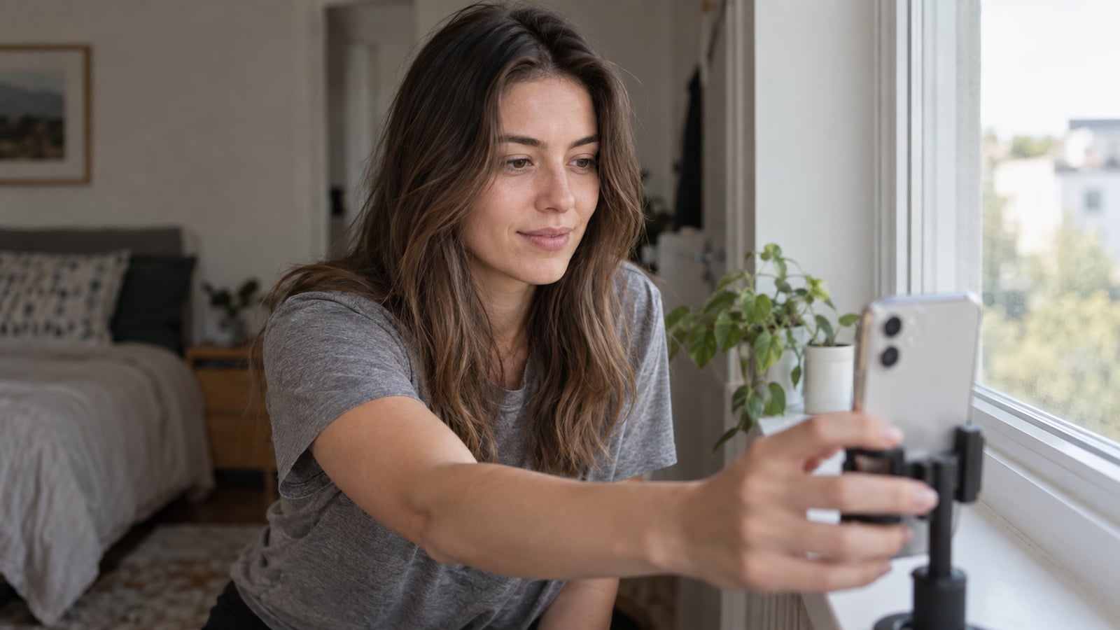 Woman in a gray shirt setting up a smartphone for a photo beside a softly lit window